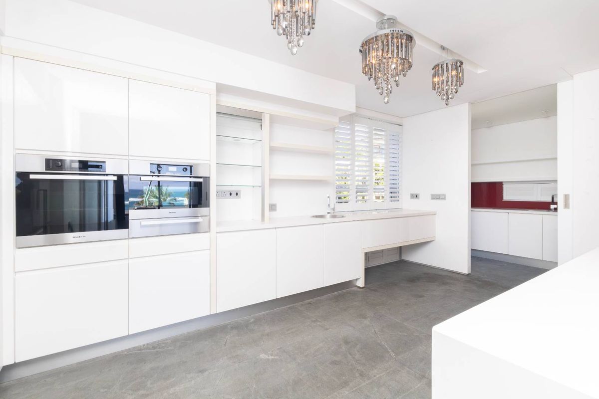 Modern, minimalist kitchen featuring high-gloss white cabinetry, built-in Miele appliances, concrete flooring, and elegant crystal chandeliers, with an adjacent scullery area.