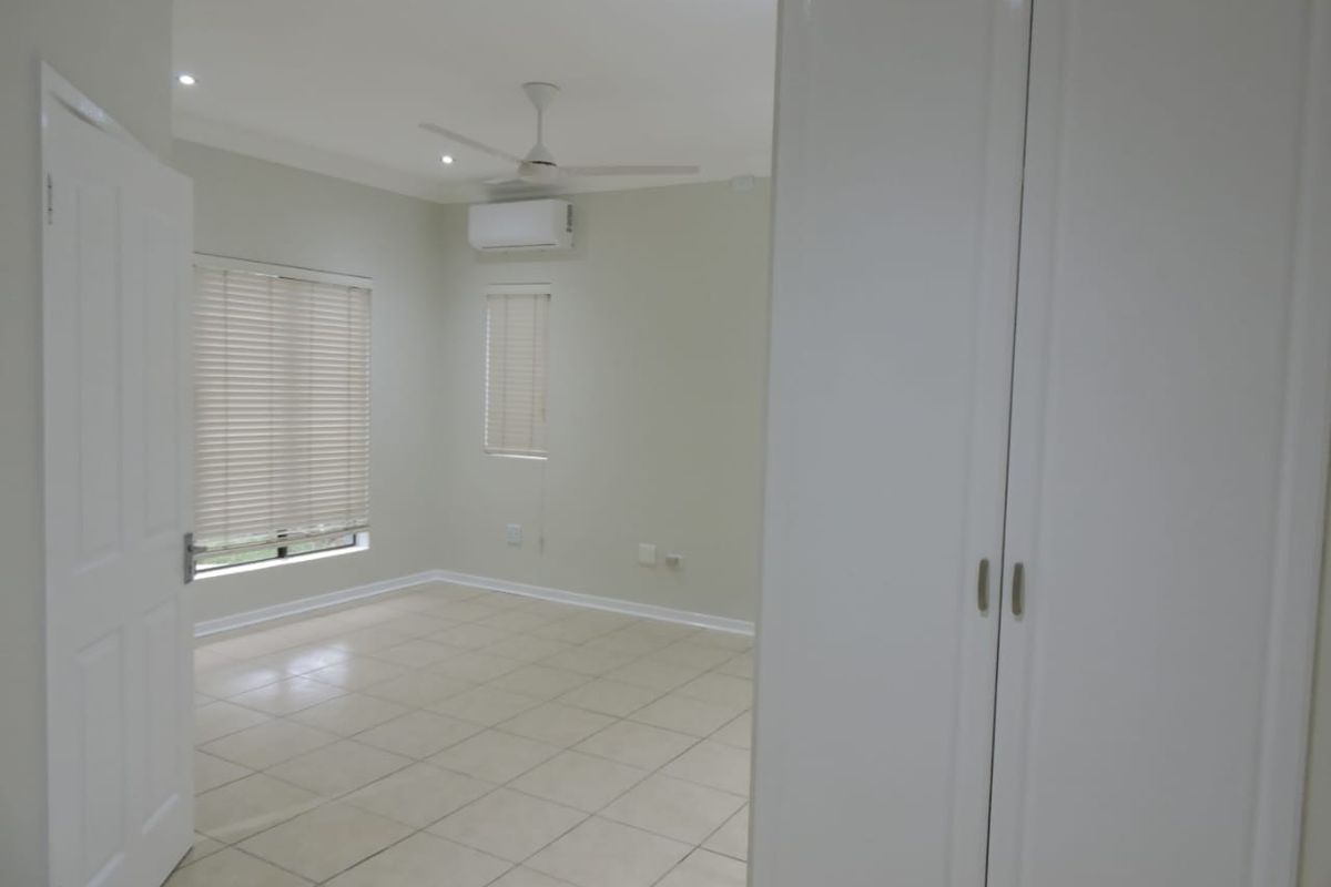 A bright, empty bedroom featuring tiled flooring, built-in cupboards, a ceiling fan, and a wall-mounted air conditioning unit.