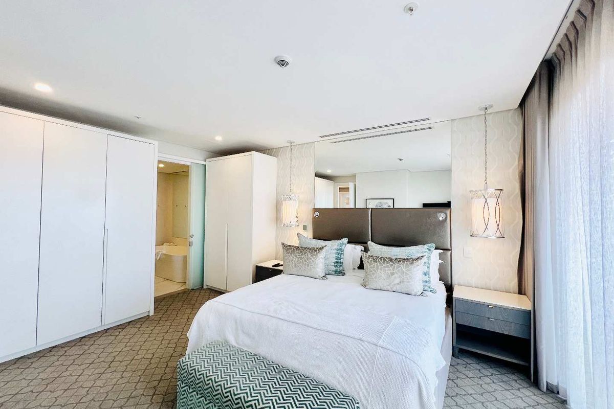 A well-appointed bedroom featuring built-in wardrobes, a mirrored feature wall, recessed lighting, and an en-suite bathroom with a glass sliding door.