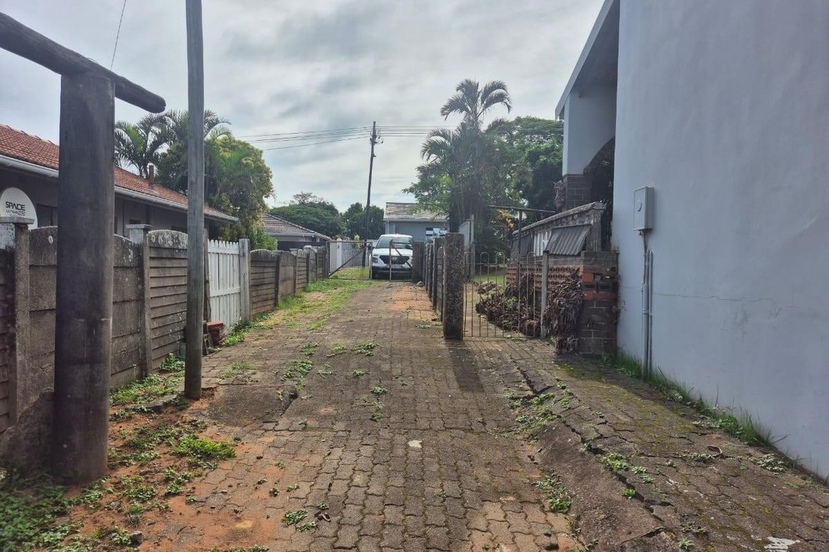 A rear exterior view showing a paved driveway area with boundary walls and metal gate access, requiring maintenance and landscaping.