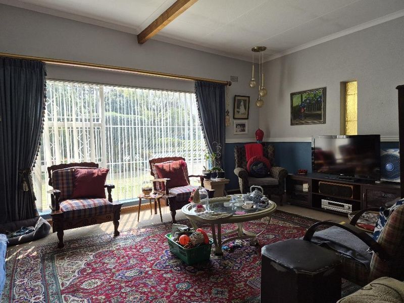 A comfortable living room featuring exposed ceiling beams, large windows with vertical blinds, and decorative pendant lighting, offering a warm and traditional atmosphere.