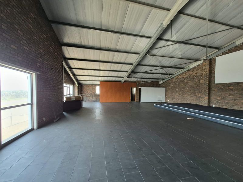 Spacious open-plan office area featuring exposed brick walls, tiled flooring, and a raised platform stage under an industrial-style exposed steel roof structure.