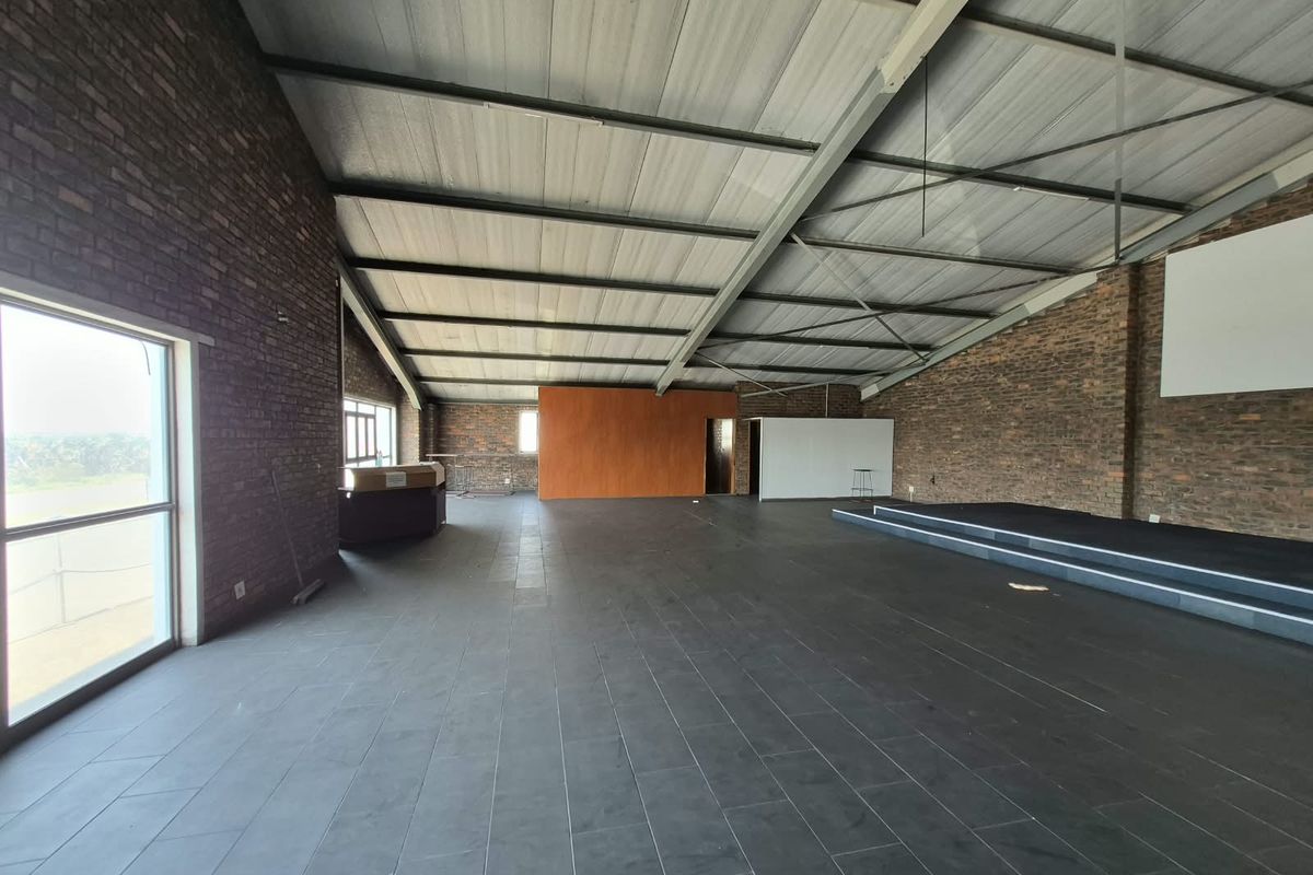 Spacious open-plan office area featuring exposed brick walls, tiled flooring, and a raised platform stage under an industrial-style exposed steel roof structure.