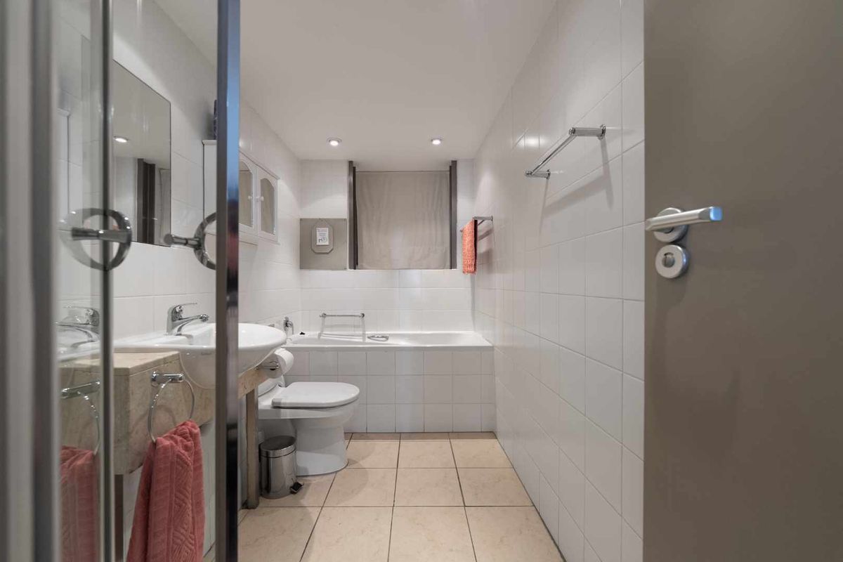 A clean, functional bathroom featuring white wall tiles, tiled flooring, a built-in bathtub, and a pedestal washbasin with modern fixtures.