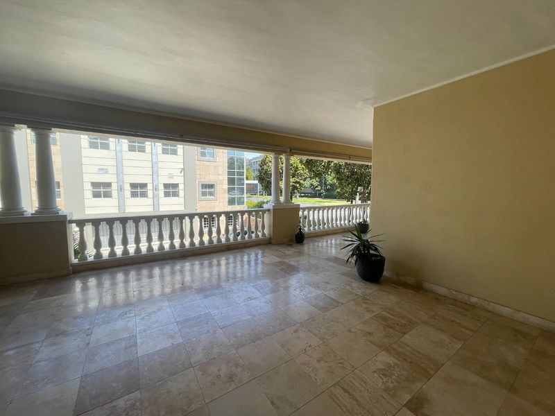 A spacious covered balcony featuring stone tile flooring, classical columns, and a decorative balustrade overlooking the surrounding area.