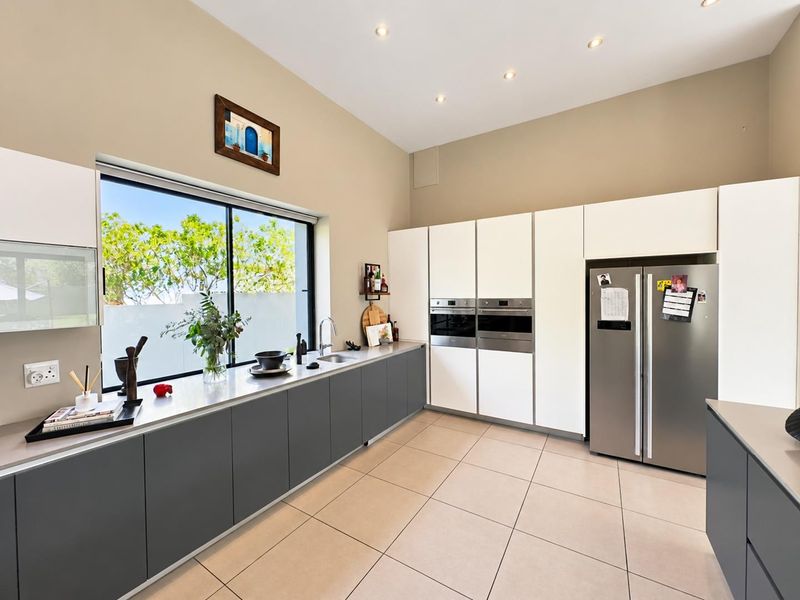 Modern kitchen featuring sleek grey and white cabinetry, stainless steel built-in ovens, stone countertops, and large windows overlooking the garden.