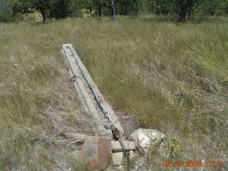 An outdoor area featuring a weathered concrete water trough with exposed metal piping set within a grassy field.