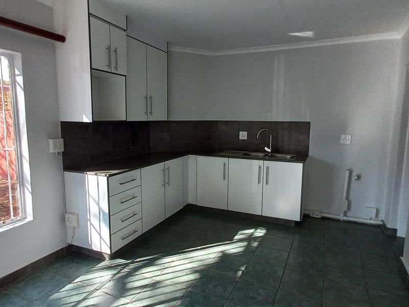 A functional, empty kitchen featuring white cabinetry, dark tiled splashbacks, and tiled flooring, with natural light entering through a large window with security bars.