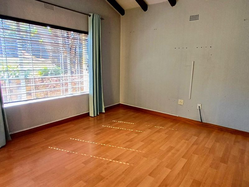 An empty bedroom featuring laminate flooring, exposed timber ceiling beams, and a large window with security bars.