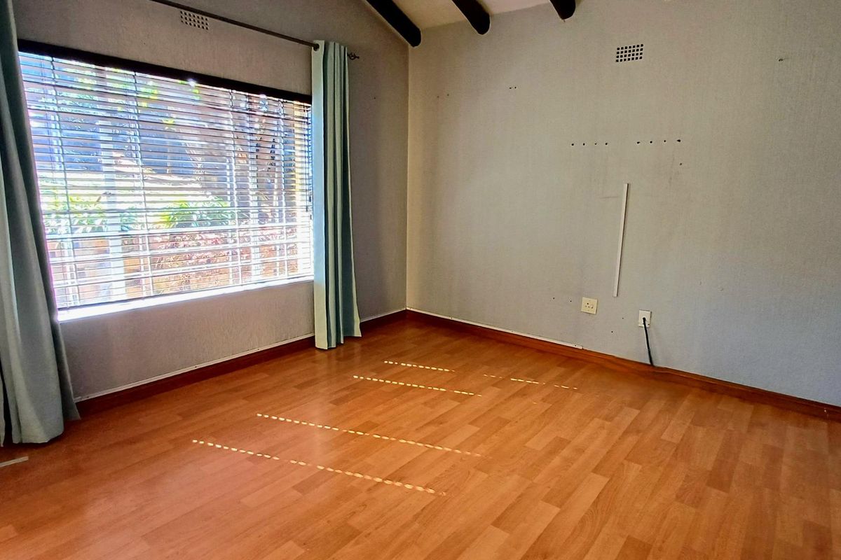 An empty bedroom featuring laminate flooring, exposed timber ceiling beams, and a large window with security bars.