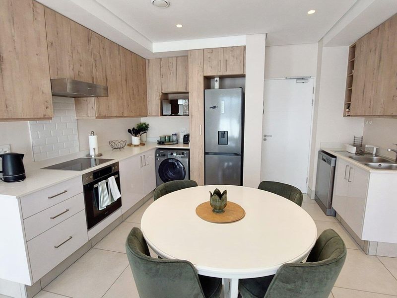 Modern open-plan kitchen featuring light wood-tone cabinetry, stone countertops, integrated appliances, and a dedicated dining area with tiled flooring throughout.