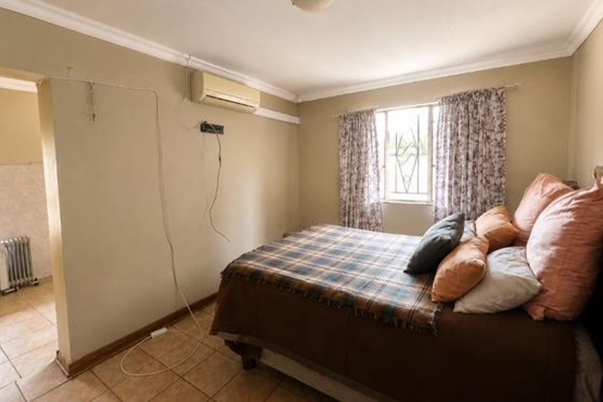 A bedroom featuring tiled flooring, security bars on the windows, and a wall-mounted air conditioning unit, with visible wall scuffs and exposed wiring.