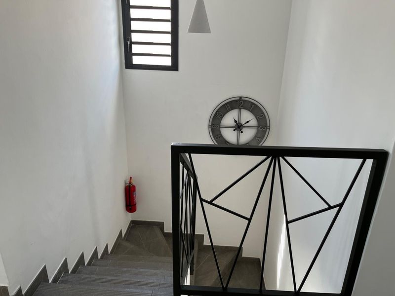 A clean, modern staircase featuring dark tiled steps and a geometric black metal balustrade, illuminated by a high-set louvered window.