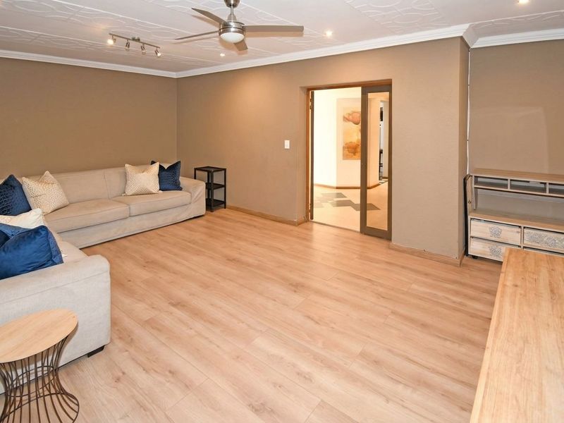 A spacious living room featuring light wood-look laminate flooring, decorative ceiling tiles, a central ceiling fan, and recessed lighting, leading to an adjacent hallway.