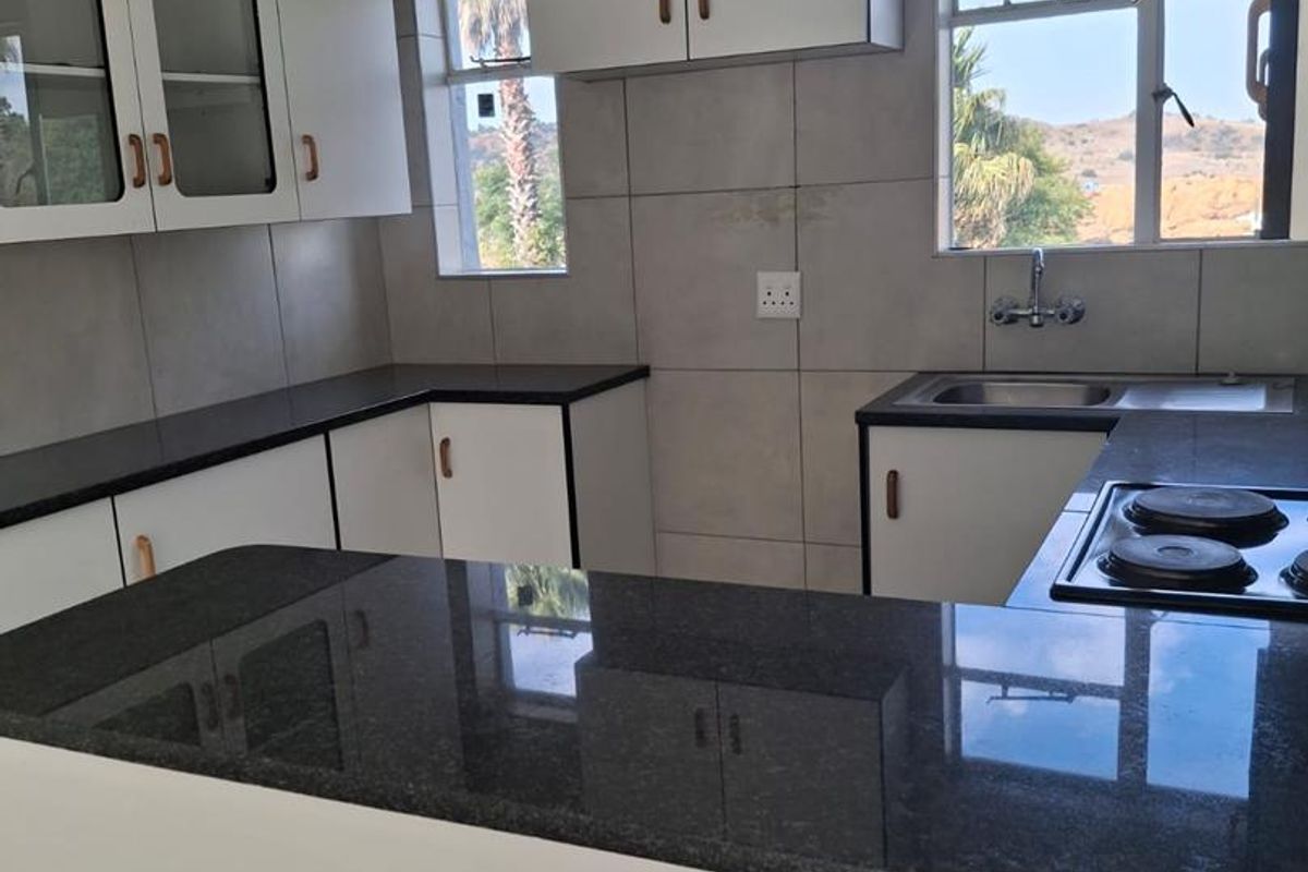 A functional kitchen featuring dark granite countertops, white cabinetry with glass-fronted upper units, a built-in electric hob, and a stainless steel sink.