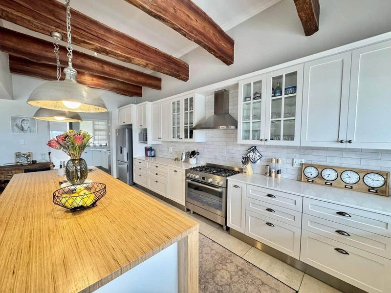 Modern farmhouse-style kitchen featuring white shaker cabinetry, stone countertops, a large timber-topped island, and striking exposed wooden ceiling beams.