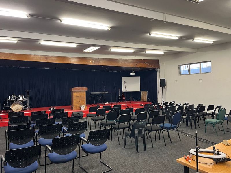 A functional multi-purpose hall featuring a raised stage area, carpeted flooring, and overhead fluorescent lighting, suitable for community gatherings or events.