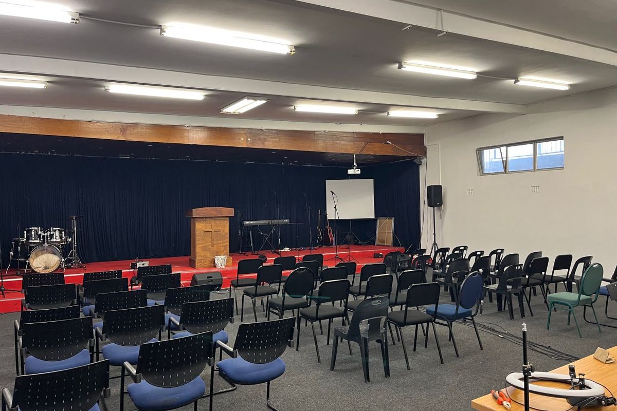 A functional multi-purpose hall featuring a raised stage area, carpeted flooring, and overhead fluorescent lighting, suitable for community gatherings or events.