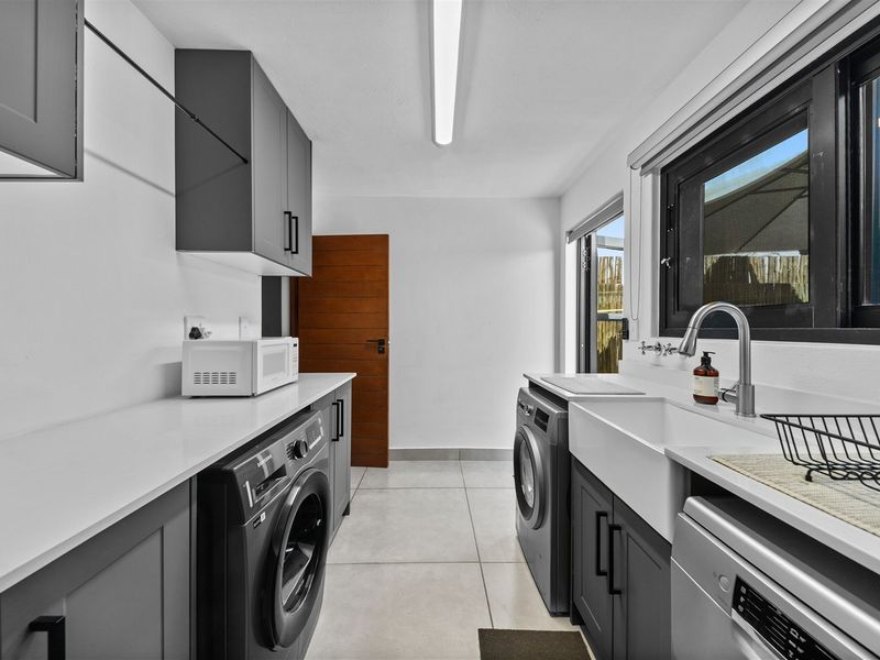 Modern, well-appointed laundry room featuring sleek grey cabinetry, quartz countertops, a farmhouse sink, and tiled flooring with direct outdoor access.