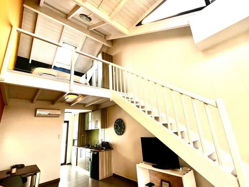 Open-plan living area featuring a mezzanine bedroom level, exposed timber ceilings, a compact kitchen, and a staircase with white railings.