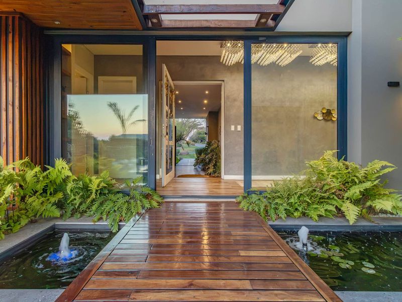 Modern entrance featuring a timber walkway over a water feature, leading through large glass sliding doors into a contemporary home interior.