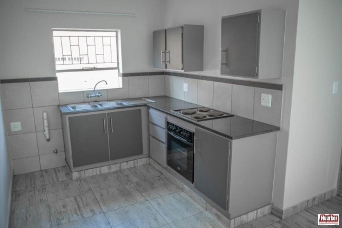 A clean, functional kitchen featuring grey built-in cabinetry, a stainless steel sink, electric hob, built-in oven, and neutral tiled finishes.