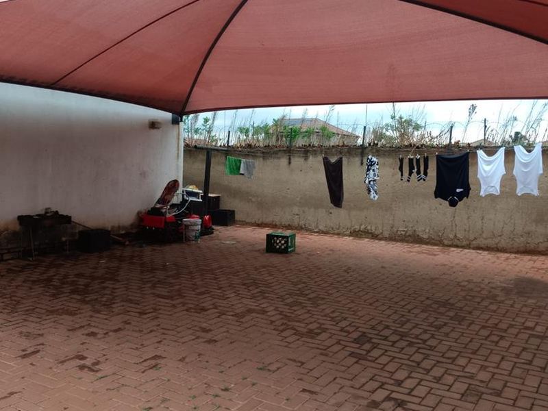A paved outdoor patio area featuring a large shade sail, enclosed by a perimeter wall with security fencing.