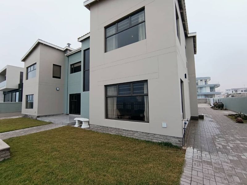 A modern two-story residential exterior featuring clean lines, stone cladding accents, large aluminium-framed windows, and a paved walkway leading to a tiled patio area.