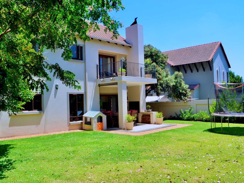 Rear view of a two-story house featuring a covered patio with a built-in braai, an upper-level balcony, and a well-maintained garden.