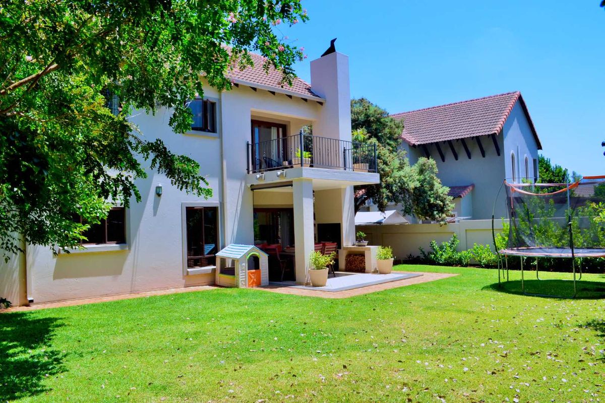 Rear view of a two-story house featuring a covered patio with a built-in braai, an upper-level balcony, and a well-maintained garden.
