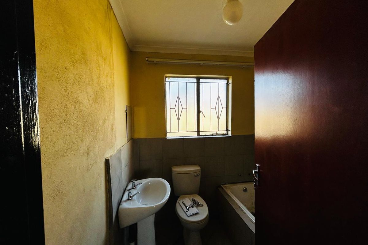 A dated bathroom featuring a pedestal basin, toilet, and bathtub, with security bars on the window and worn wall finishes.