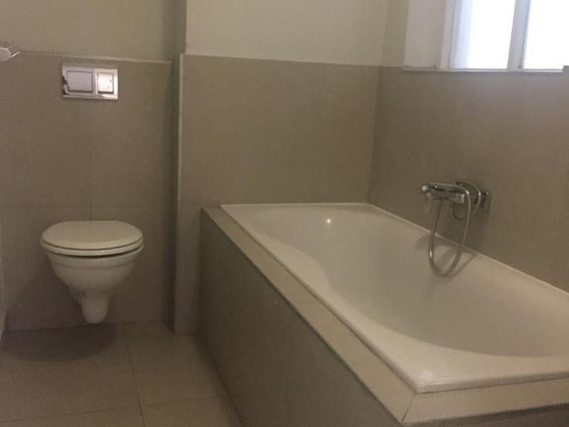 A clean, minimalist bathroom featuring a built-in bathtub, wall-mounted toilet, and neutral tiled finishes throughout.