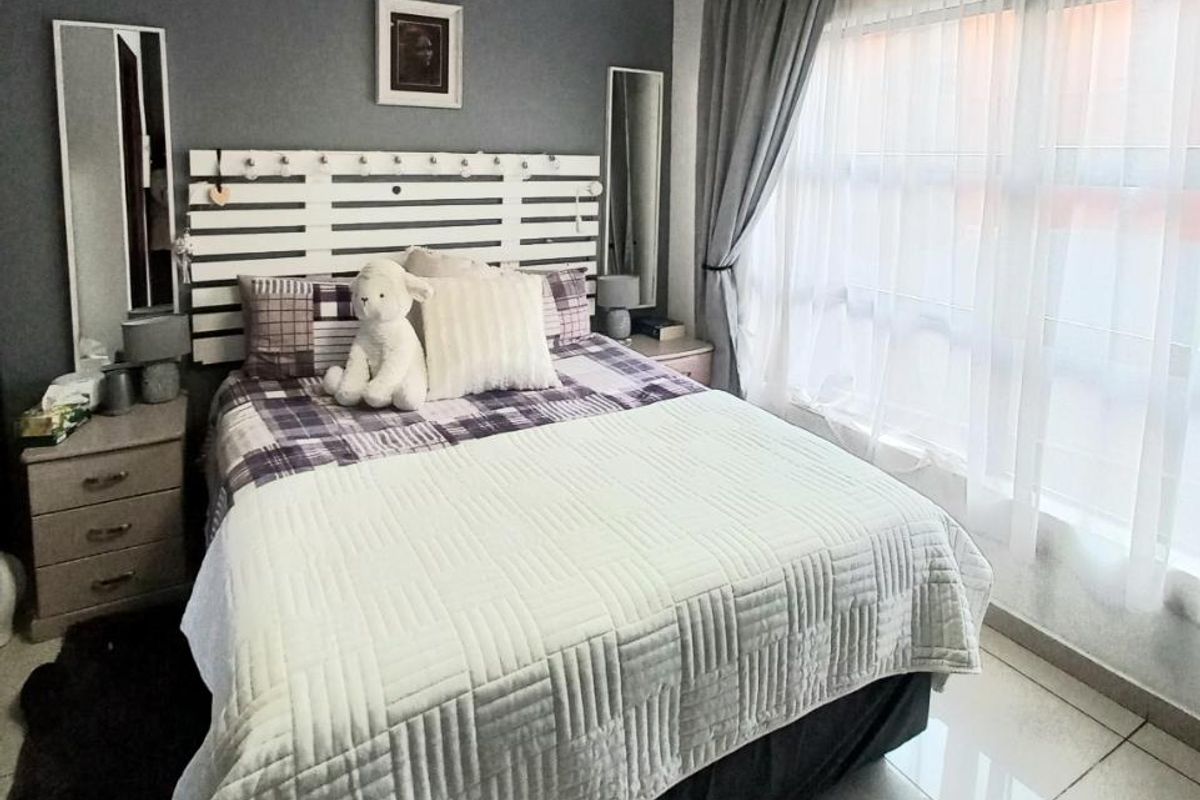 A well-maintained bedroom featuring large windows with sheer curtains, tiled flooring, and a neutral grey accent wall.
