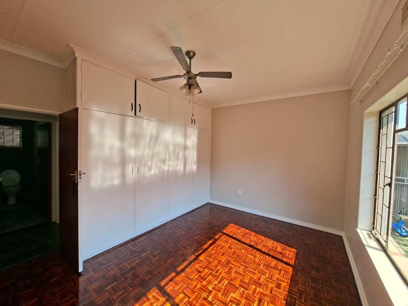 A bright, empty bedroom featuring classic parquet flooring, extensive built-in cupboard storage, and a ceiling fan.