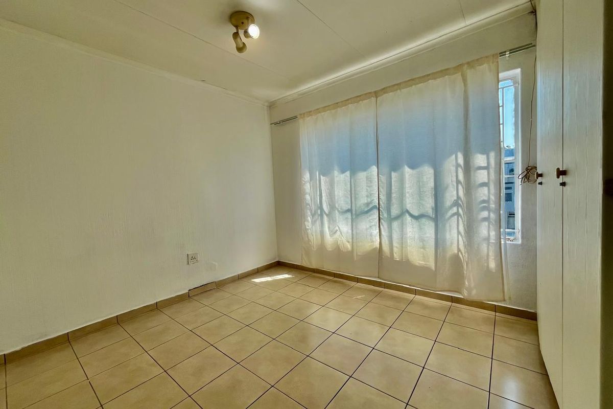A bright, empty bedroom featuring neutral floor tiles, built-in cupboards, and a window with security bars.