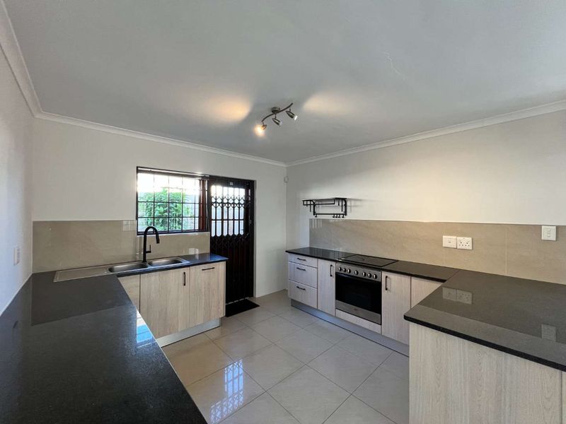 Modern kitchen featuring light wood-finish cabinetry, dark granite countertops, built-in oven and hob, and tiled flooring with direct access to the exterior.