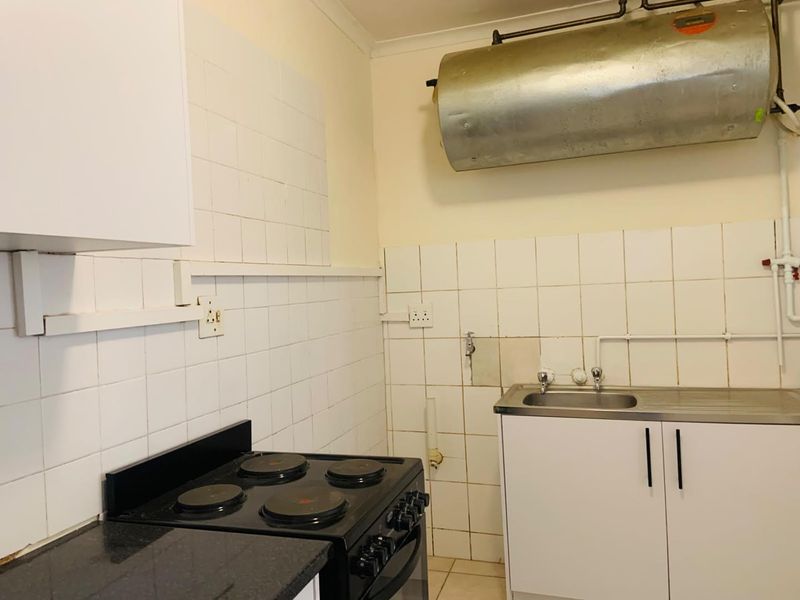 A functional kitchen area featuring a basic electric stove, stainless steel sink, and tiled splashback, with visible exposed plumbing and water heater infrastructure.