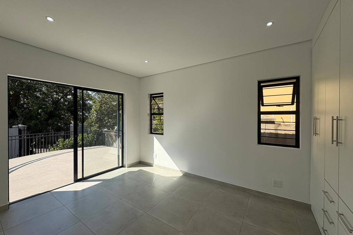 A bright, modern bedroom featuring neutral tiled flooring, built-in cupboards, and a large sliding door opening onto an outdoor balcony.