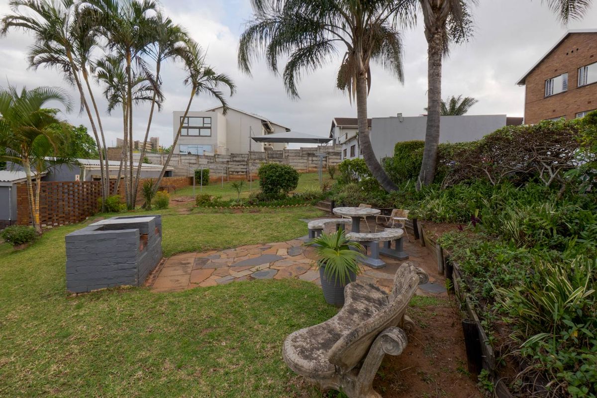 A spacious rear garden featuring a built-in brick braai, stone paved patio area, established palm trees, and tiered landscaping.