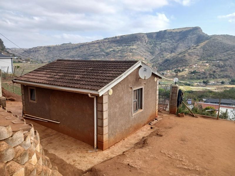 Rear view of a detached house featuring a tiled roof and satellite dish, set against a scenic mountainous landscape with an unpaved yard.
