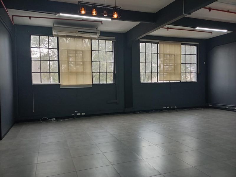 Spacious open-plan office area featuring industrial-style steel windows, exposed ceiling beams, tiled flooring, and modern pendant lighting.