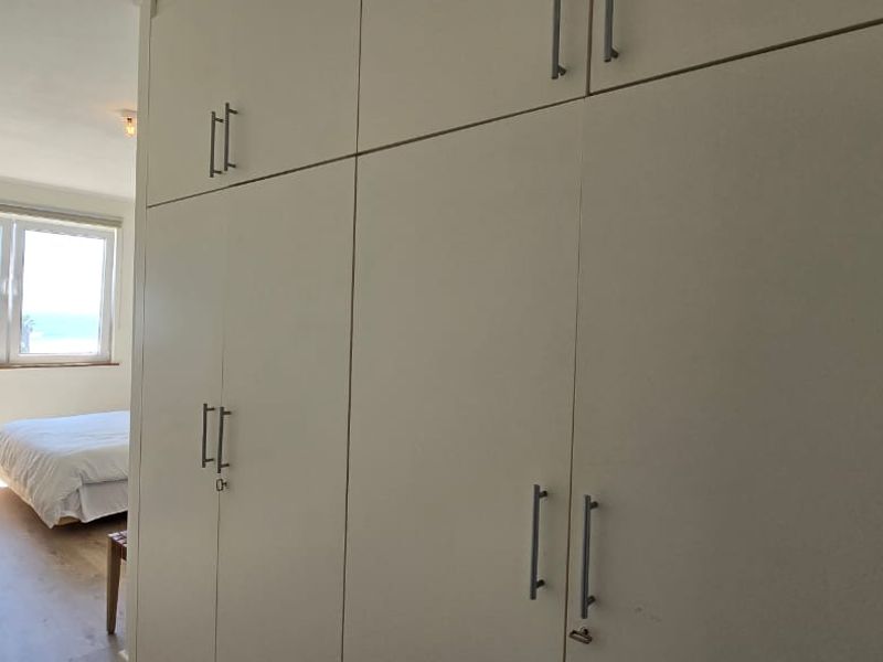 A bright bedroom featuring extensive built-in cupboards, light laminate flooring, and a window offering a scenic ocean view.