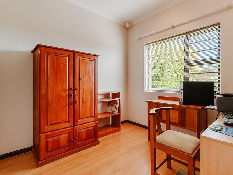 A functional bedroom or home office space featuring light-toned laminate flooring and a window fitted with venetian blinds.