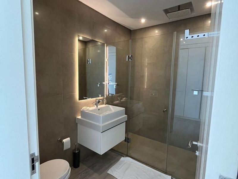 Modern bathroom featuring a wall-mounted vanity, backlit mirror, glass-enclosed rain shower, and wall-hung toilet with contemporary tiled finishes.