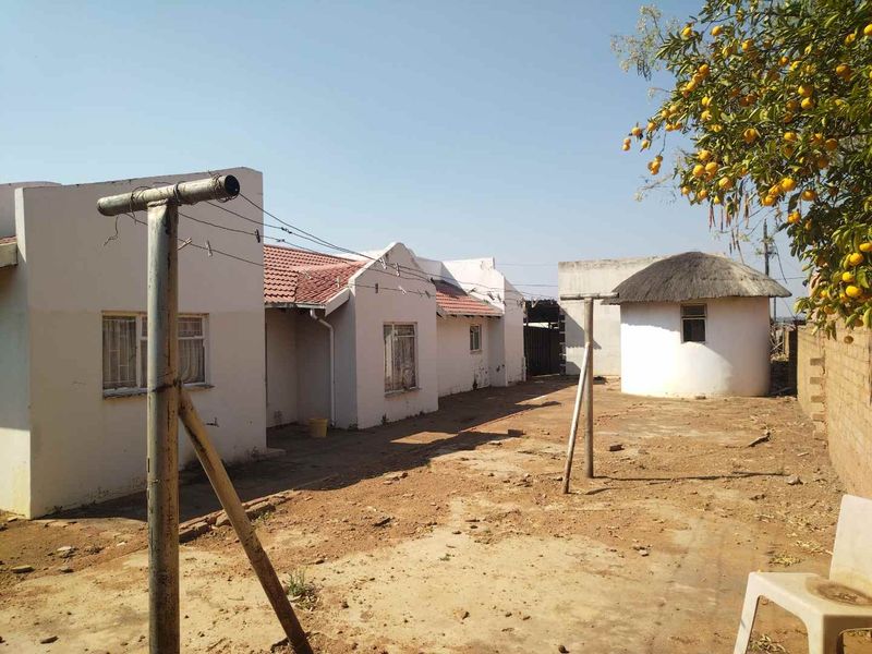 Rear view of the property featuring a main house with tiled roof, a separate thatched rondavel, and an unpaved yard area.