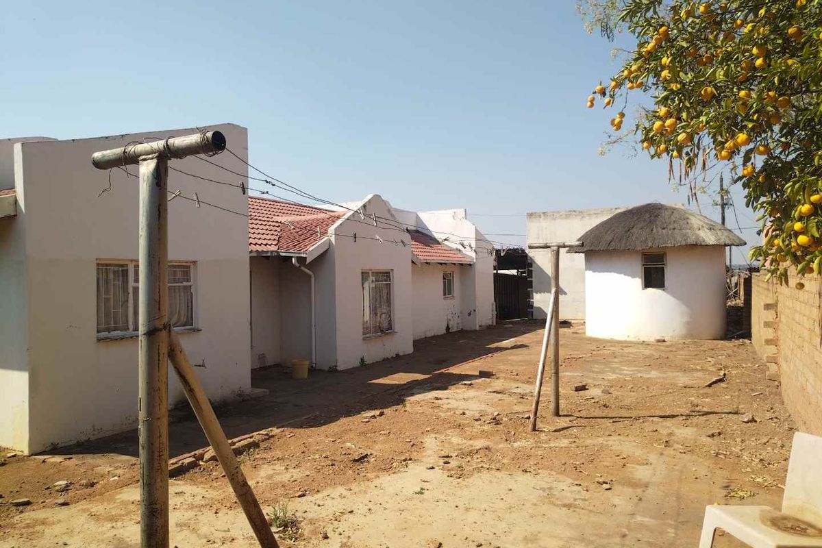 Rear view of the property featuring a main house with tiled roof, a separate thatched rondavel, and an unpaved yard area.