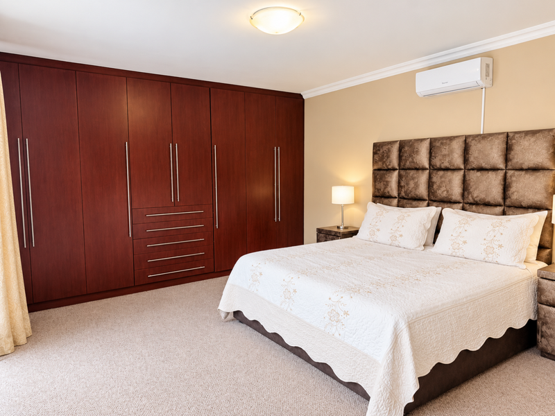 Spacious bedroom featuring extensive dark wood built-in cupboards, wall-to-wall carpeting, and a wall-mounted air conditioning unit.
