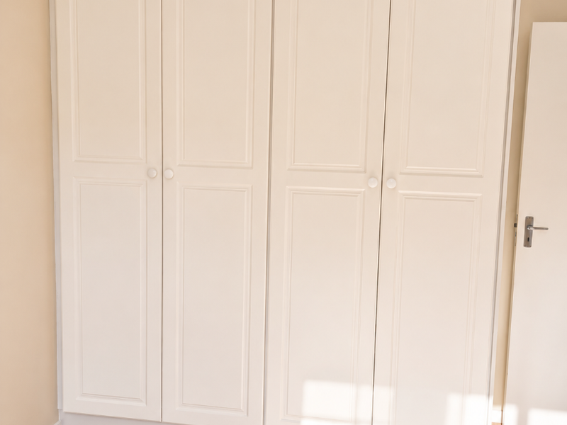 A bright bedroom featuring neutral walls, light wood-look laminate flooring, and a large, well-maintained built-in wardrobe with classic panel detailing.