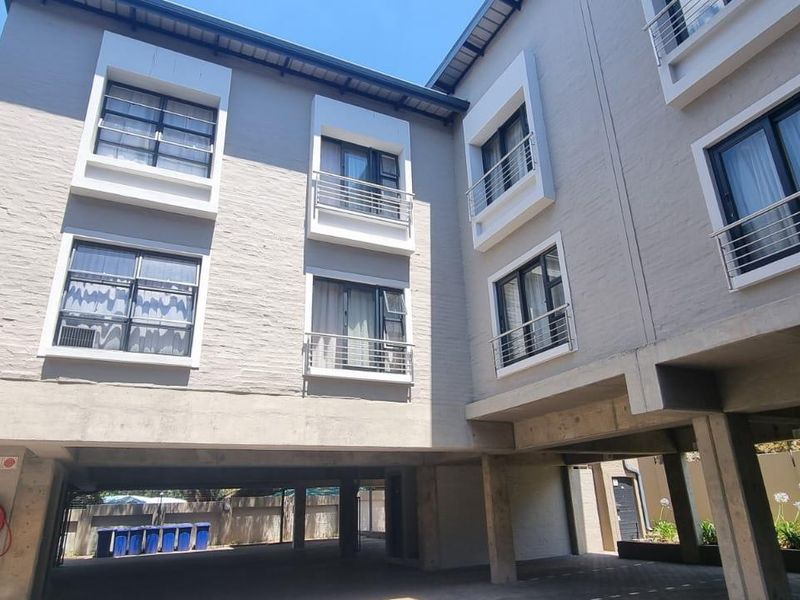 Exterior view of a modern apartment building featuring concrete structural pillars, metal balustrades, and aluminium-framed windows with a paved ground level.