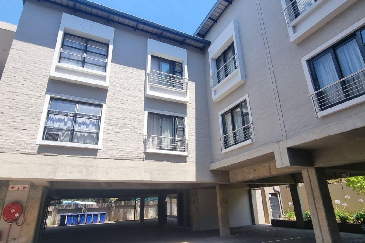 Exterior view of a modern apartment building featuring concrete structural pillars, metal balustrades, and aluminium-framed windows with a paved ground level.